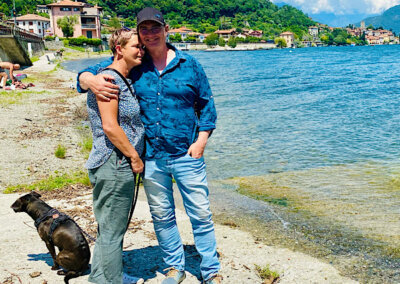 Eng umschlungen stehen Freunde am Wasser. Der Hund schaut in eine andere Richtung. Blauer Himmel und Sonnenschein.